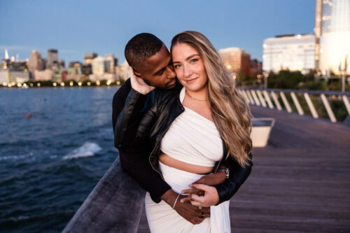 Engaged couple embraces and smiles on a boardwalk by the water in Hudson River Park at sunset during their New York City engagement session