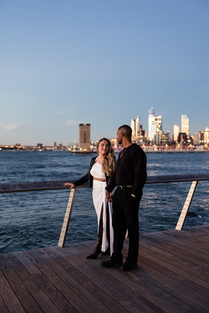 Engaged couple stands on a wooden deck by the water in Hudson River Park, with the New York City skyline glowing in the sunset behind them