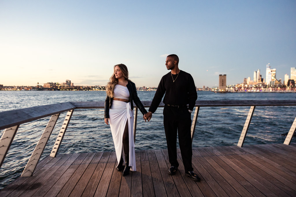 A couple holding hands on a boardwalk by the water with a city skyline in the background at sunset, capturing a romantic NYC engagement moment.