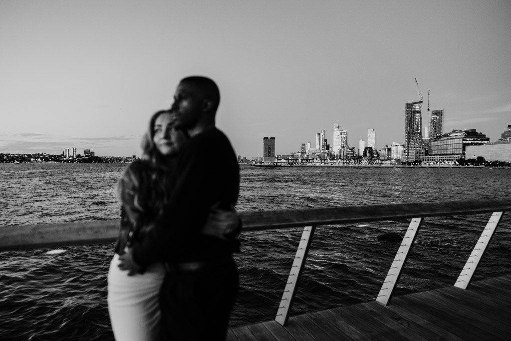 Creative black and white deep focus photo of engaged couple embracing by the water, New York City skyline in the distance