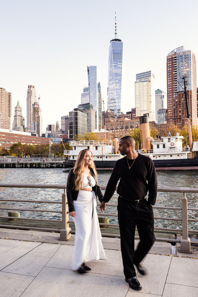 A couple holding hands walks through Hudson River Park with New York City skyscrapers and a boat in the background