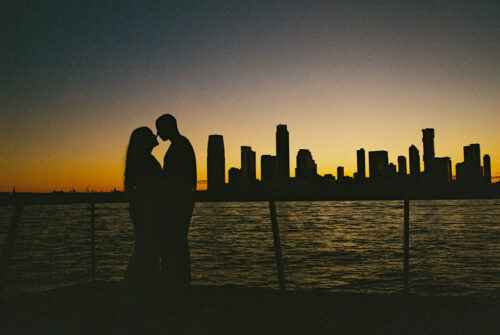 Silhouetted couple embraces by the water at sunset, with New York Coty skyline in the background during fall engagement session