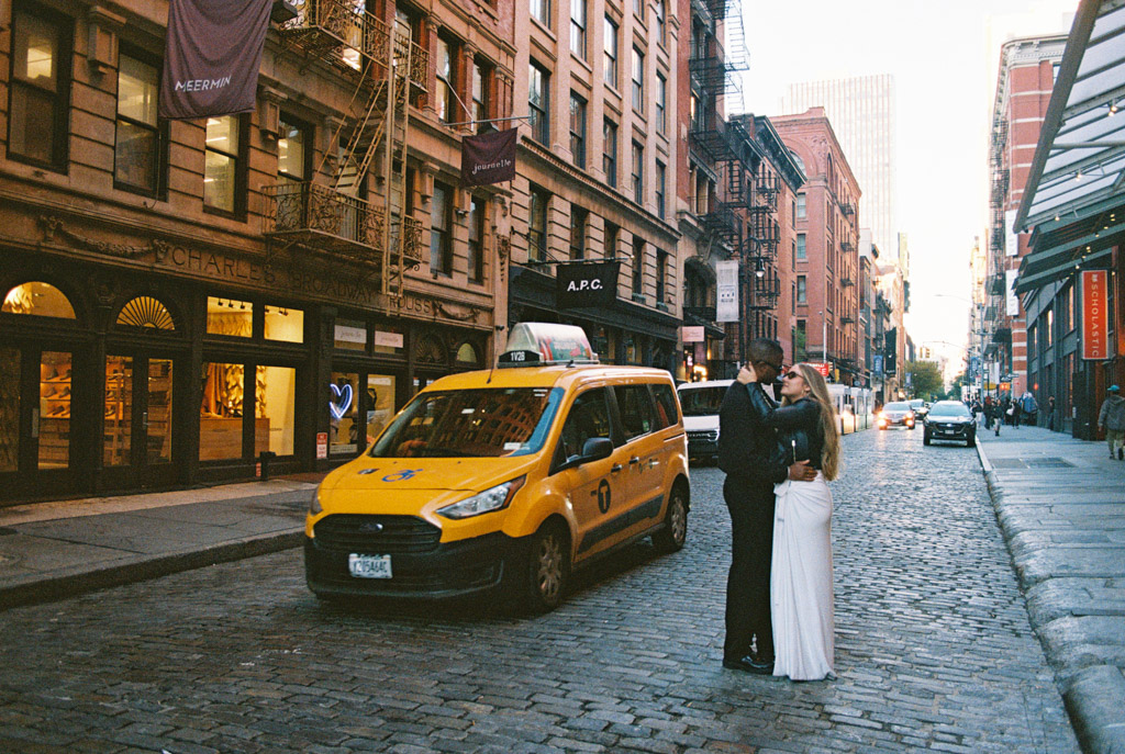 Engaged couple embraces on a cobblestone street next to a yellow taxi during New York City engagement session