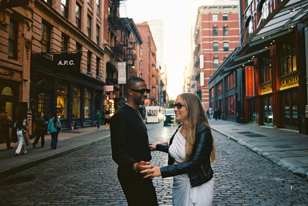 A couple smiles and holds hands on a cobblestone street during their New York City engagement sessiojn, surrounded by city buildings