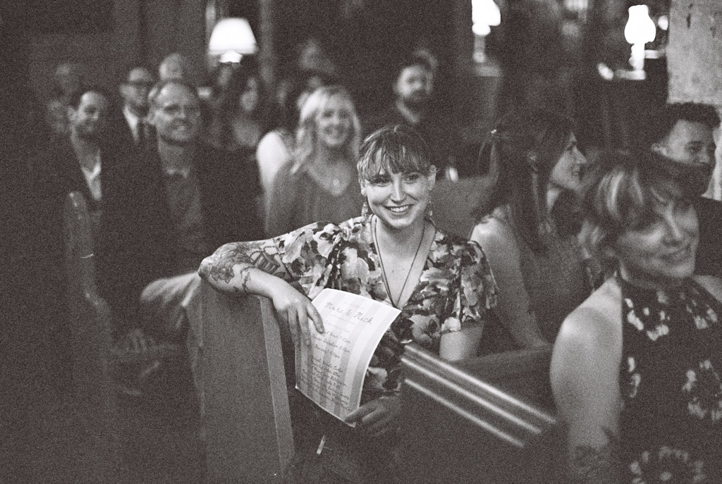 Black and white film photo of woman smiling and holding a program while seated among others at a Salvage One wedding ceremony
