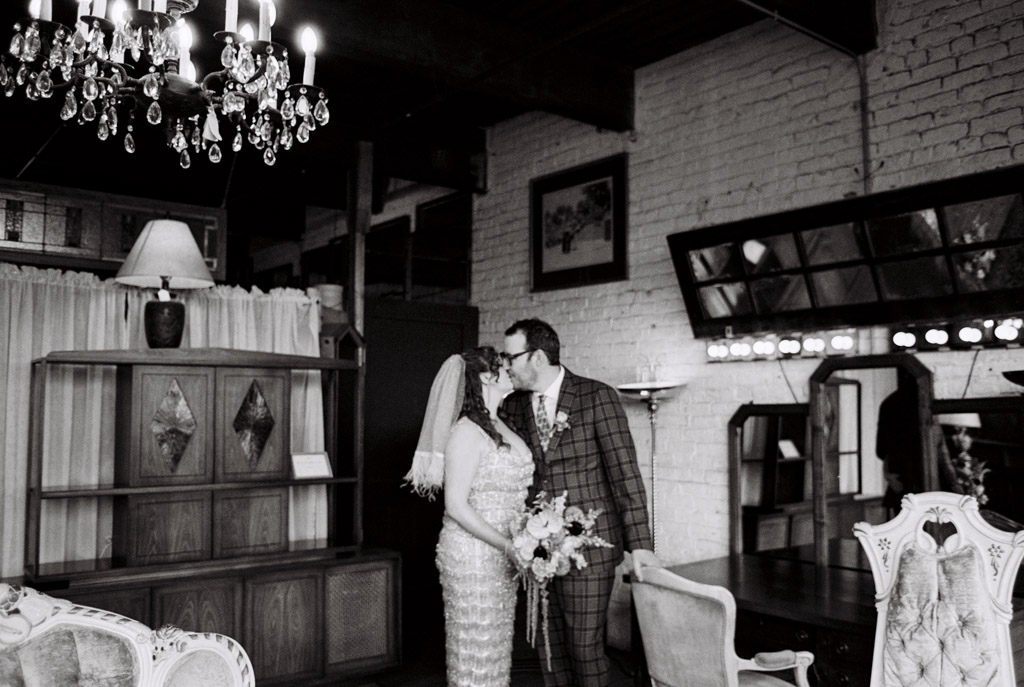 Black and white film photo of bride and groom kissing in a room filled with antique furniture and chandeliers inside Salvage One wedding venue