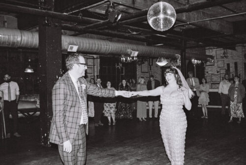 A couple dances under a disco ball as guests watch in an industrial-style venue, capturing the unique charm of a Salvage One wedding.