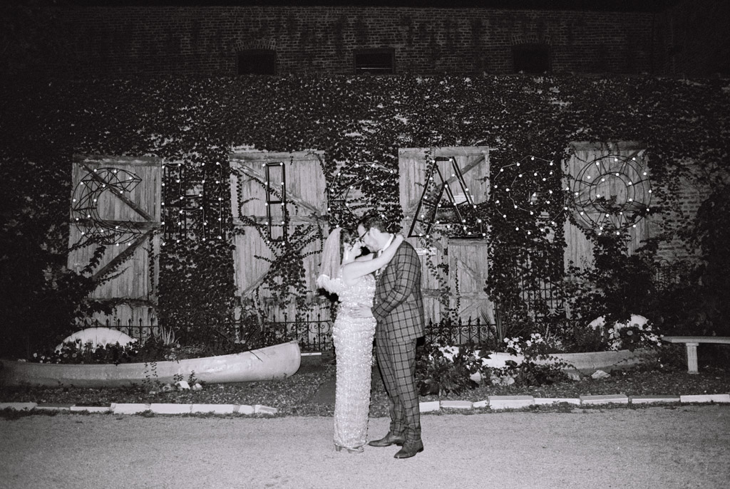 Black and white photo of bride and groom embracing at night in front of a vine-covered wall outside Salvage One