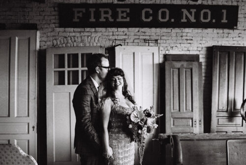 A couple stands smiling with a bouquet in front of vintage doors and a “FIRE CO. NO.1” sign—capturing the charm of a Salvage One wedding.