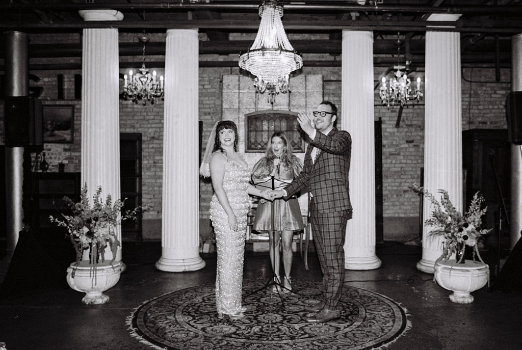Black and white film photo of bride and groom standing with an officiant under a chandelier during their Salvage One wedding ceremony in a beautifully decorated hall