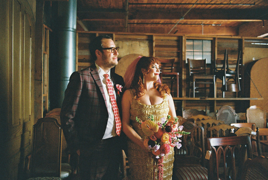 Film photo of bride and groom standing together inside Salvage One, surrounded by wooden chairs, with the bride holding a colorful bouquet