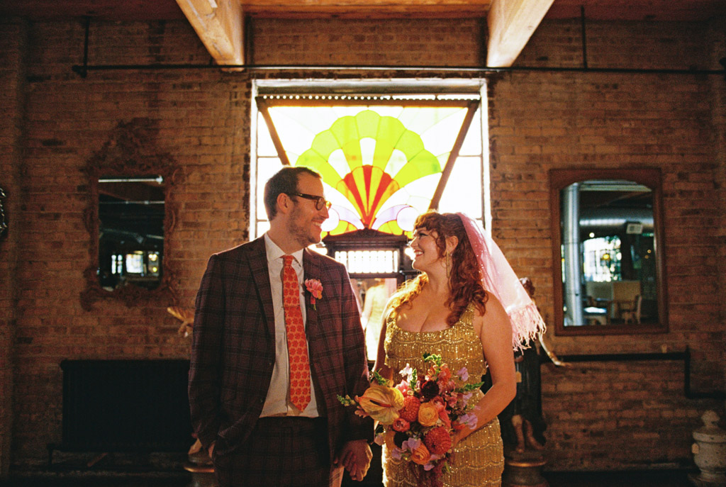 Film photo of smiling couple in wedding attire standing indoors with a colorful stained glass window behind them inside Salvage One in Chicago