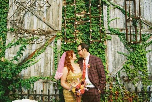 A bride and groom smile at each other in front of an ivy-covered rustic wooden wall at their Salvage One wedding.