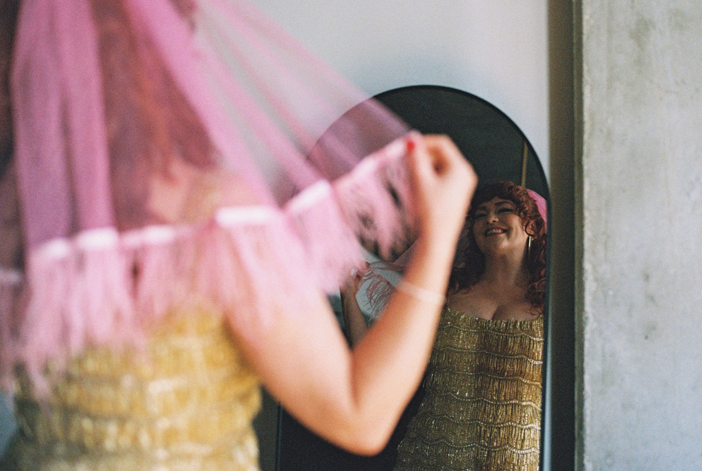 Film photo of bride in a gold dress and pink veil smiing at her reflection in a mirror before her Salvage One wedding