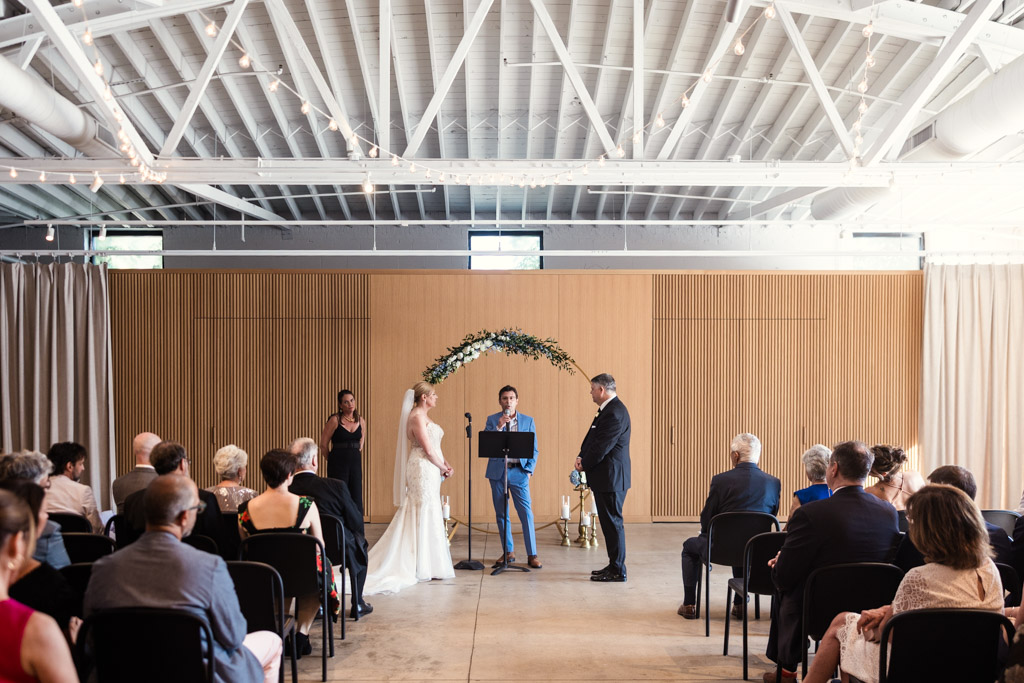 A bride and groom stand at the altar indoors, surrounded by seated guests during their wedding ceremony at Sarabande Chicago