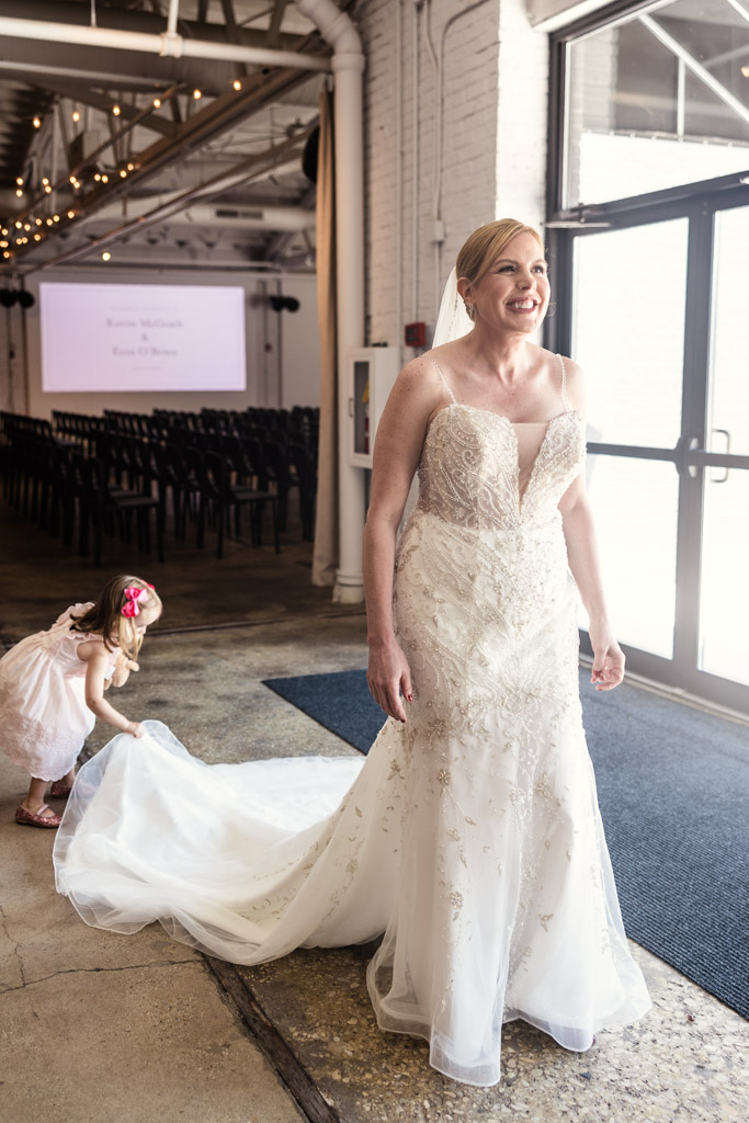 Bride in a white wedding dress smiles as a young girl adjusts her train near large windows inside Sarabande Chicago