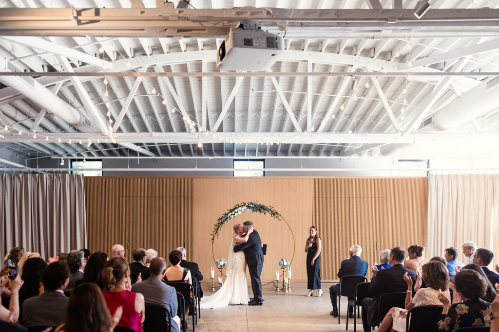 Newlyweds kiss under a floral arch after their indoor wedding ceremony inside Sarabande Chicago, as guests watch and applaud