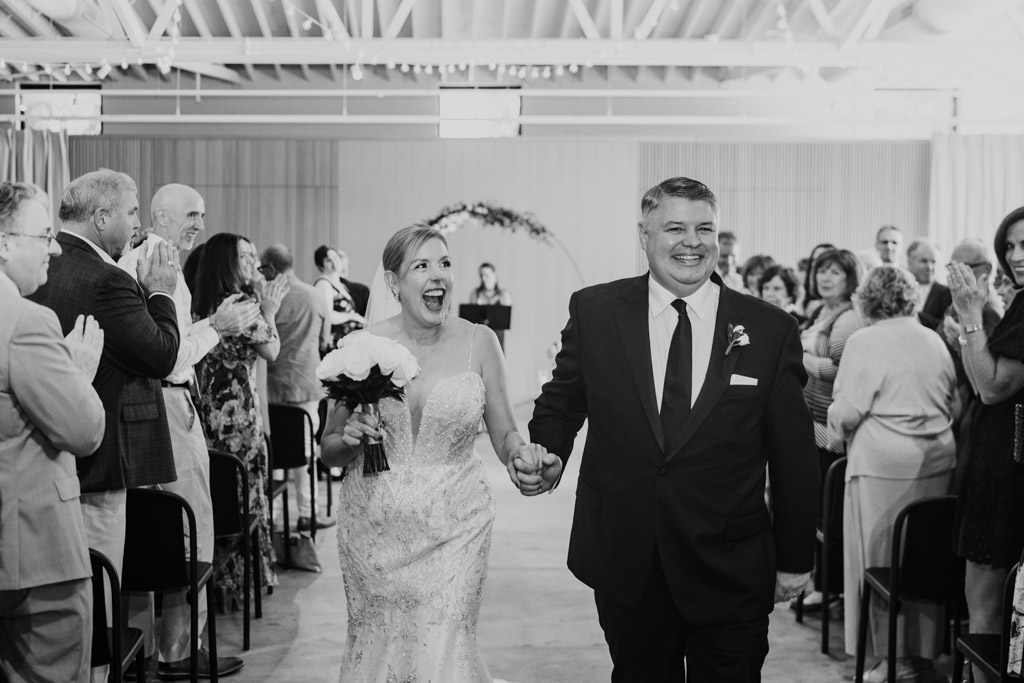 A newlywed couple smiles and holds hands while walking down the aisle to a gentle sarabande as guests applaud.