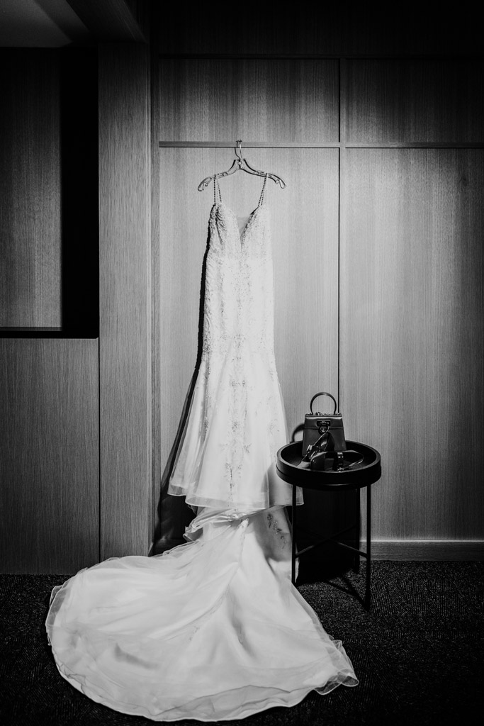 Black and white photo of bride's dress and train hanging on a wall next to a small table with her purse and shoes for wedding celebration at Sarabande Chicago