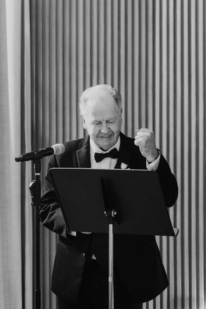 Black and white photo of man in a tuxedo speaking at a podium with a microphone during Sarabande Chicago wedding celebration