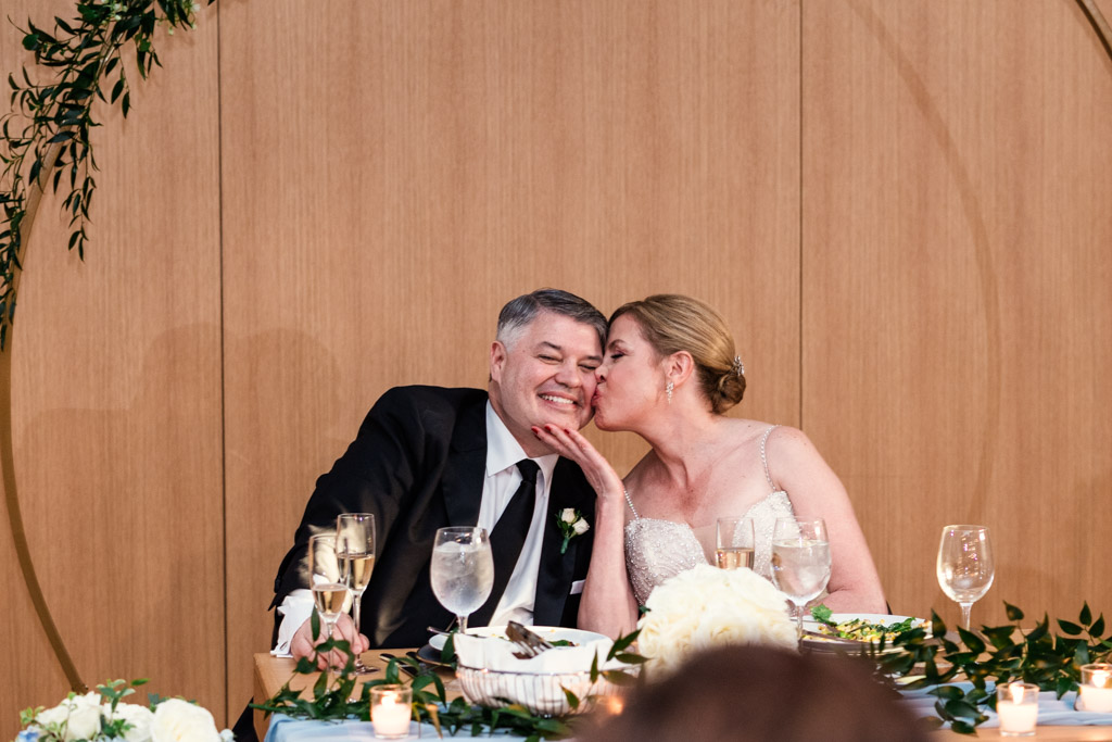 Bride kisses groom's cheek as they smile, sitting at a sweetheart table with flowers and glasses during their wedding reception at Sarabande Chicago