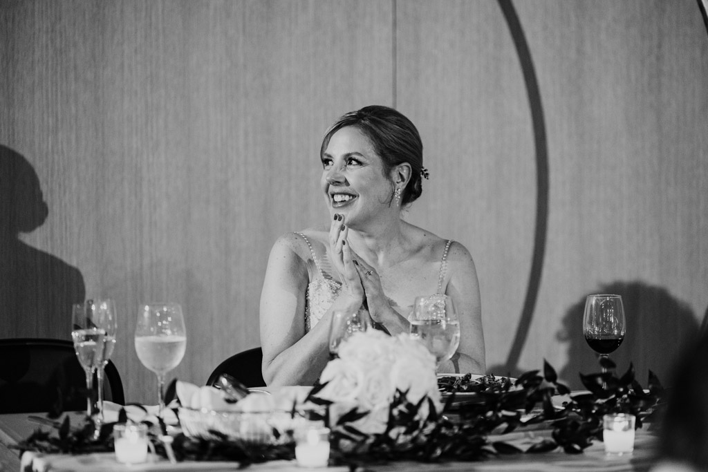 Black and white photo of bride smiling and clapping seated at the sweetheart table with candles and wine glasses during her Sarabande Chicago wedding reception