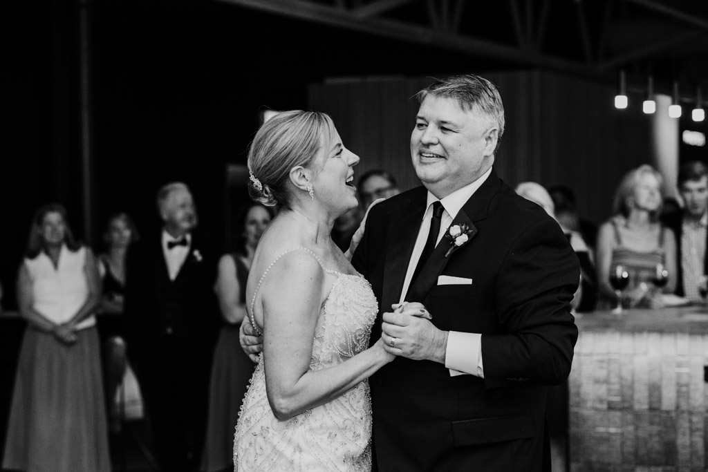 A couple in formal attire smile and dance a graceful Sarabande at a celebration, with guests watching in the background.