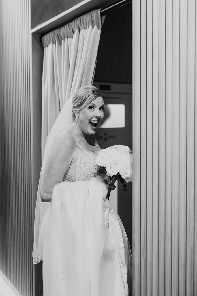 Black and white photo ofsmiling bride holding a bouquet and her dress train stands by a doorway with curtains