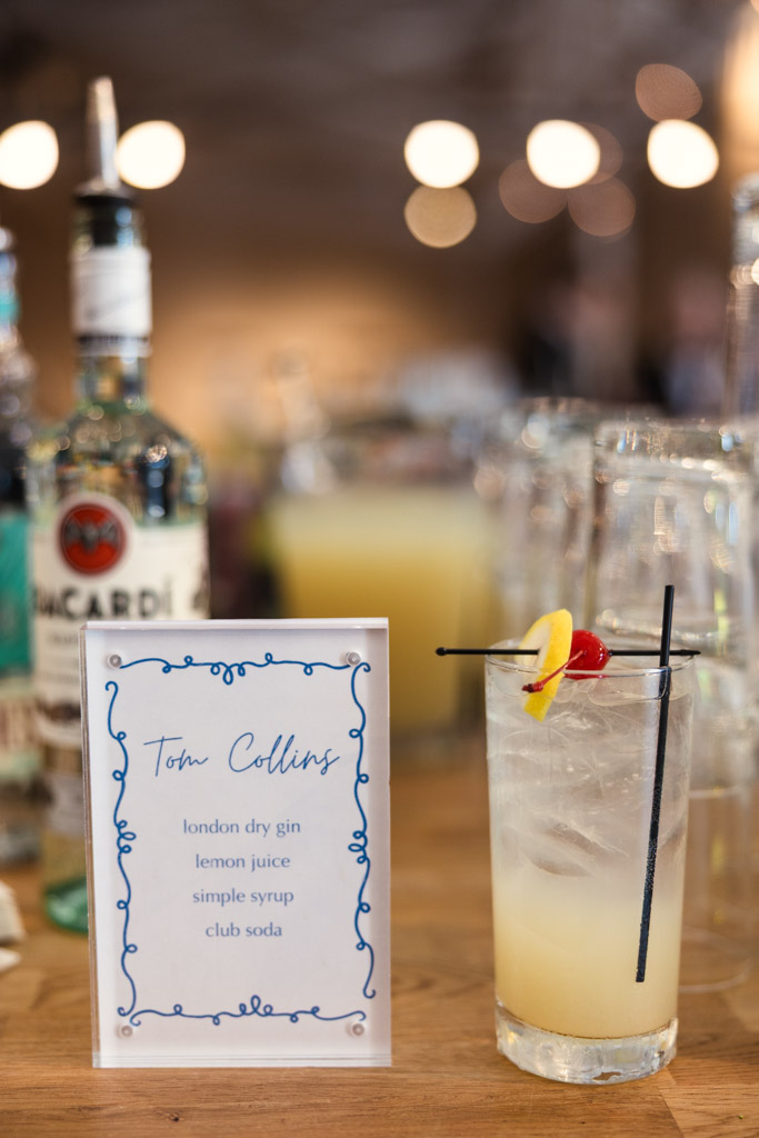A Tom Collins cocktail with lemon and cherry garnish sits beside a recipe card and bottles on a bar counter for wedding reception at Sarabande Chicago