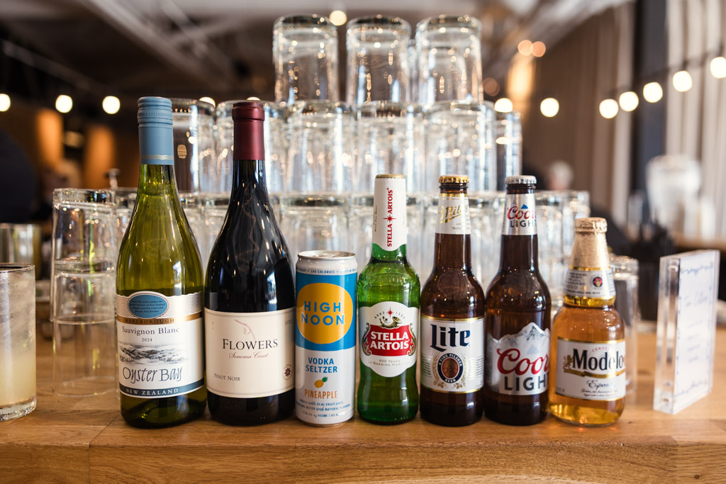 Bottles and cans of various alcoholic beverages lined up on a wooden bar with stacked glasses behind them for wedding reception at Sarabande Chicago