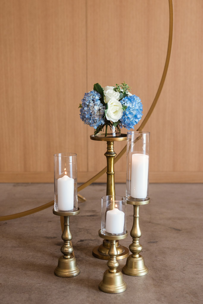 Blue and white flowers in a vase with three lit candles on brass stands create a serene sarabande of light and color against the wooden background for summer wedding celebration at Sarabande Chicago