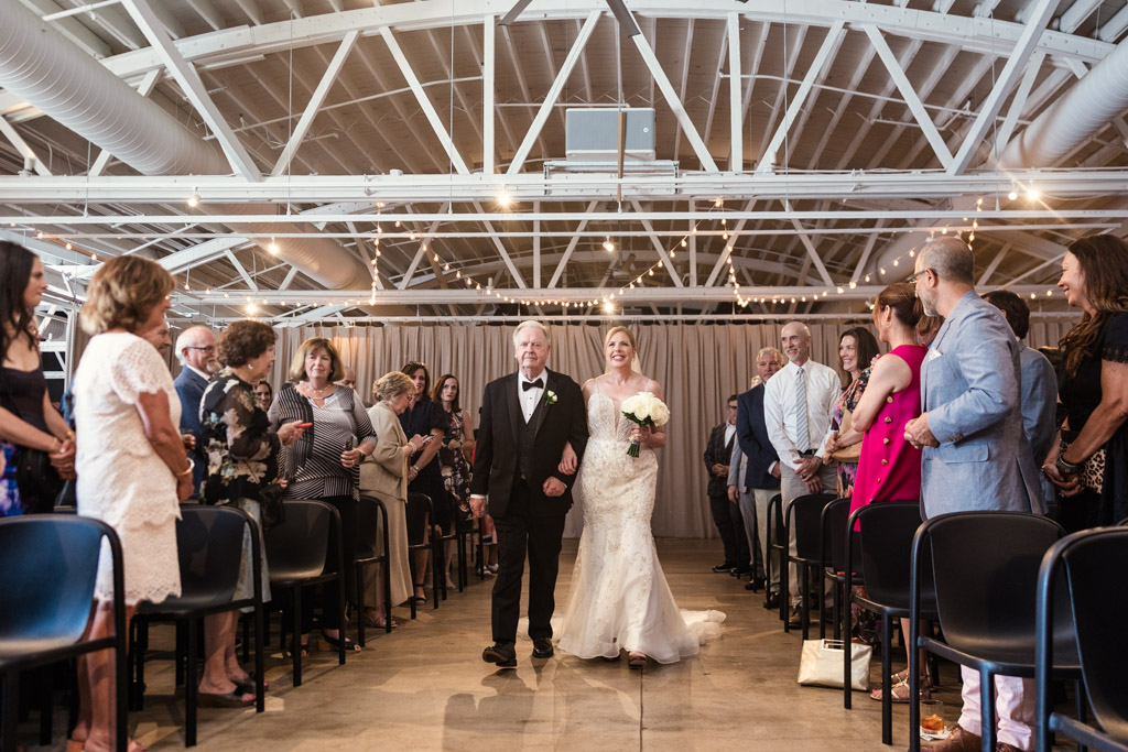 A bride in a white dress walks down the aisle with her father as guests watch and smile during summer wedding ceremony at Sarabande Chicago