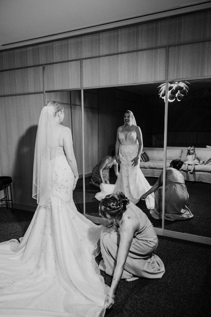 Black and white photo of bride in a wedding dress standing in front of a mirror as another woman adjusts her gown