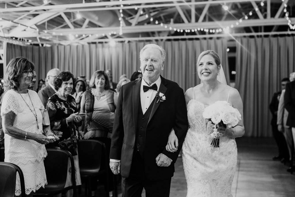 Black and white photo of happy bride walking down the aisle with her father in a tuxedo during wedding ceremony at Sarabande Chicago