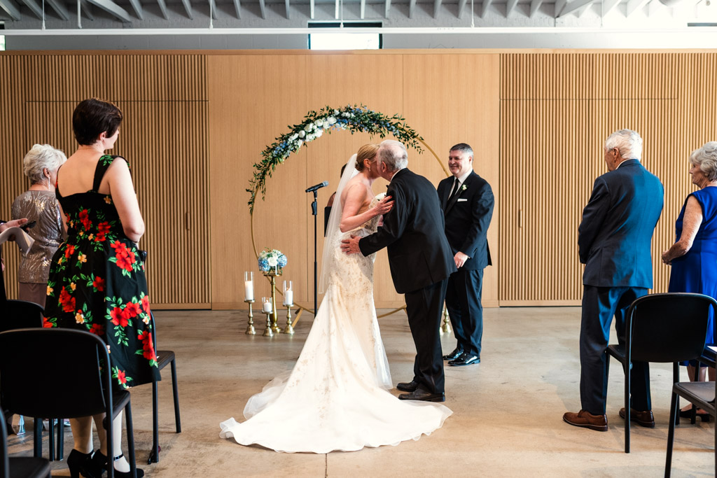 A bride in a white dress is kissed on the cheek by her father during an indoor wedding ceremony at Sarabande Chicago