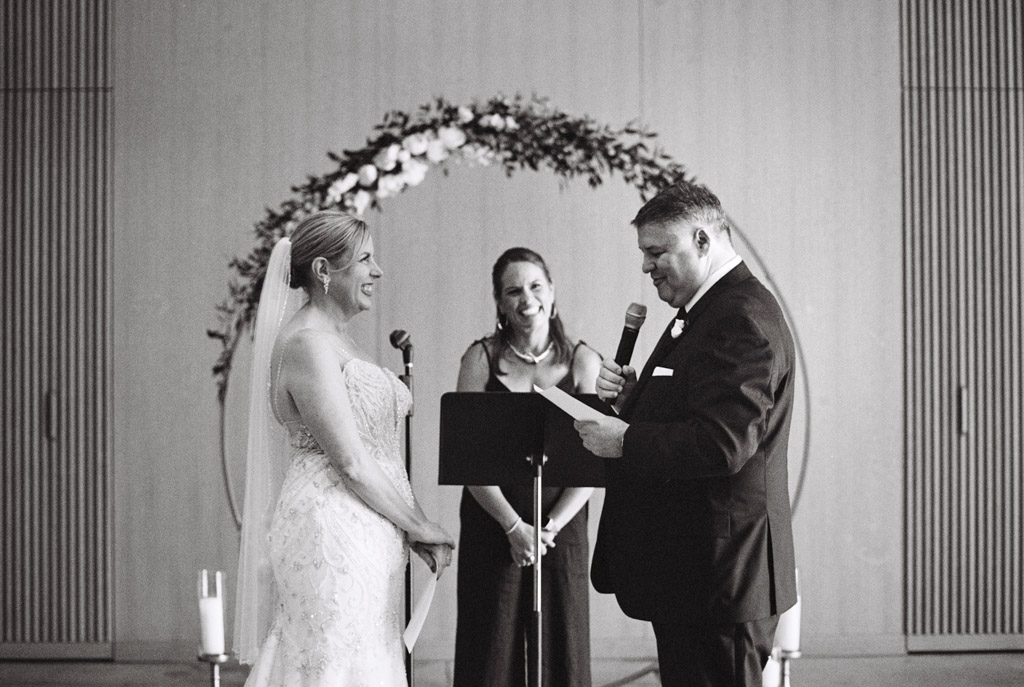 Black and white photo of groom reading vows to bride, smiling, with an officiant and a floral arch in the background during wedding ceremony at Sarabande Chicago
