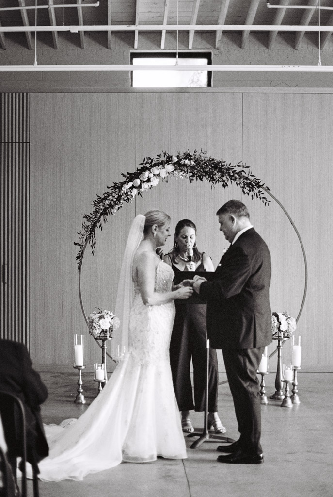 Black and white photo of bride and groom exchanging rings during their indoor wedding ceremony before an officiant and floral arch at Sarabande Chicago