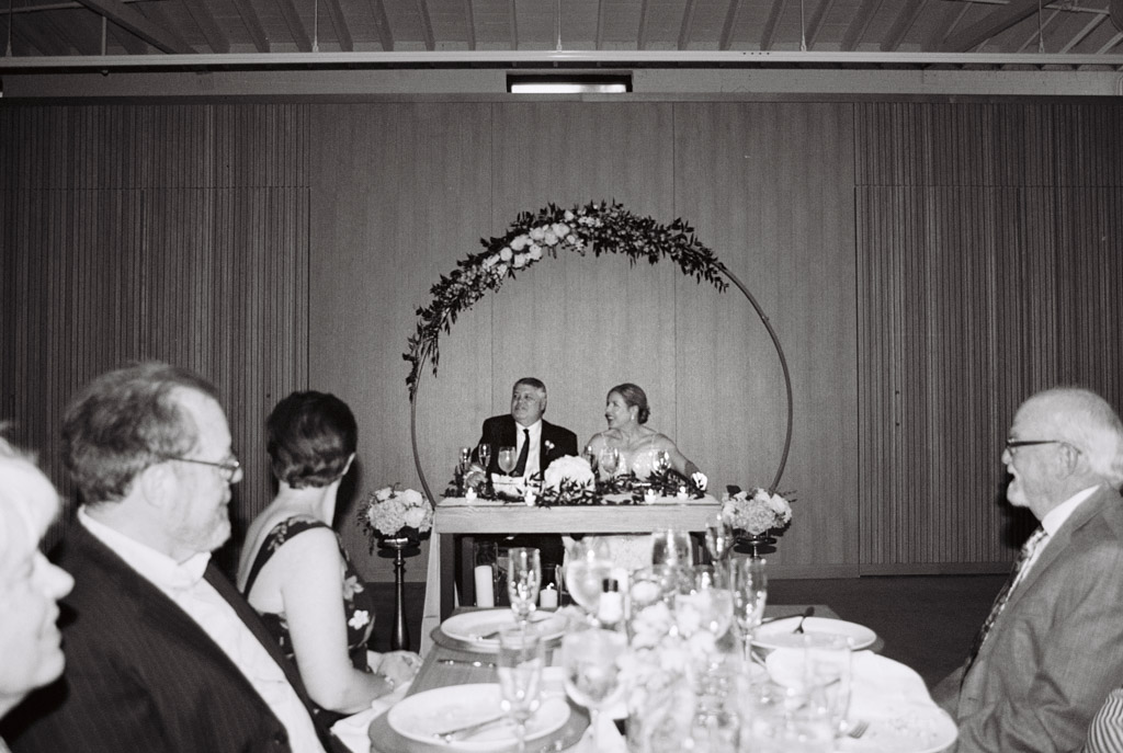 Black and white photo of bride and groom seated at a decorated table, facing wedding guests inside Sarabande Chicago