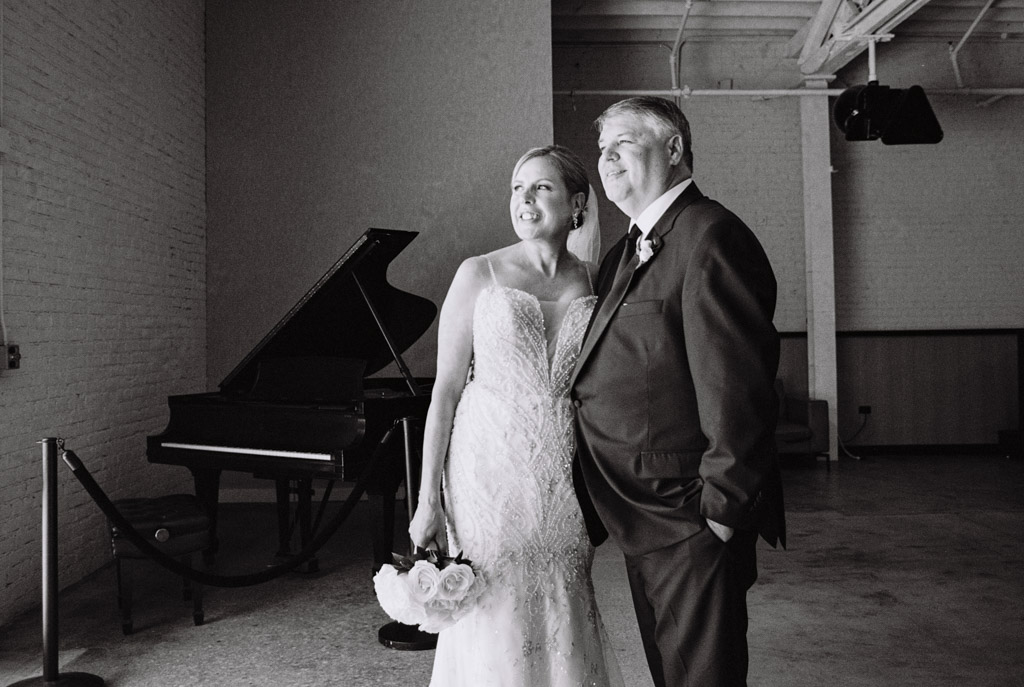 Black and white photo of bride and groom standing together indoors, smiling, with a grand piano in the background inside Sarabande Chicago