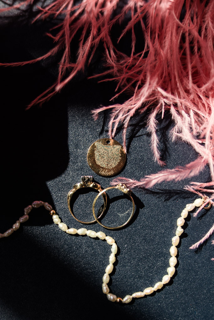 Gold rings, a pendant, and a pearl necklace rest on a dark surface with pink feathers in the background for Salvage One wedding