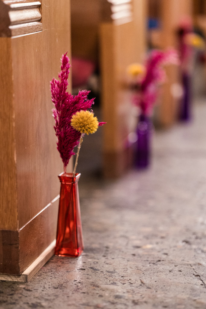 Red vase with dried pink and yellow flowers placed on the floor beside a wooden pew in a row inside Salvage One wedding ceremony space