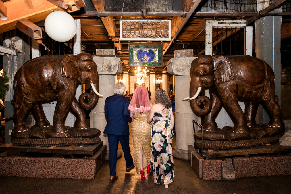 A bride walks with her parents between large carved elephant statues in the wedding ceremony space at Salvage One