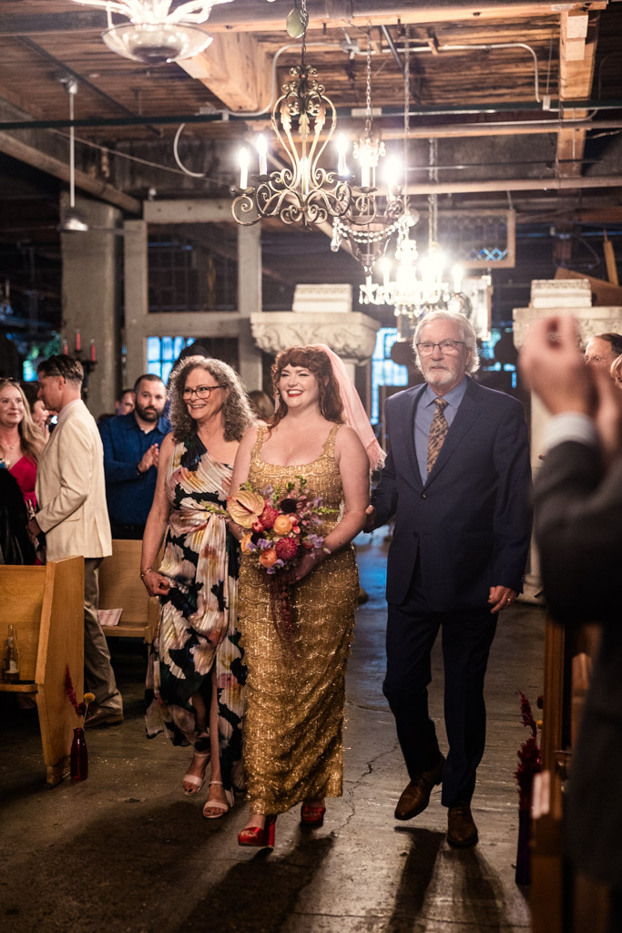 A smiling bride in a gold dress walks down the aisle with her parents at her side during. her Salvage One wedding
