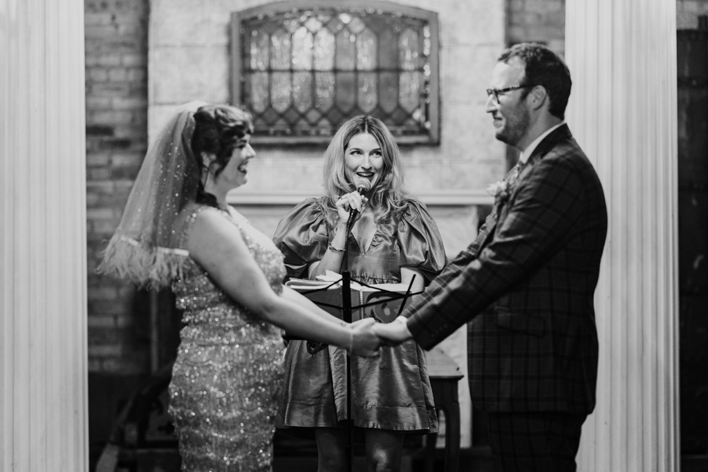 Black and white photo of bride and groom holding hands, smiling, while the officiant speaks during their Salvage One wedding ceremony