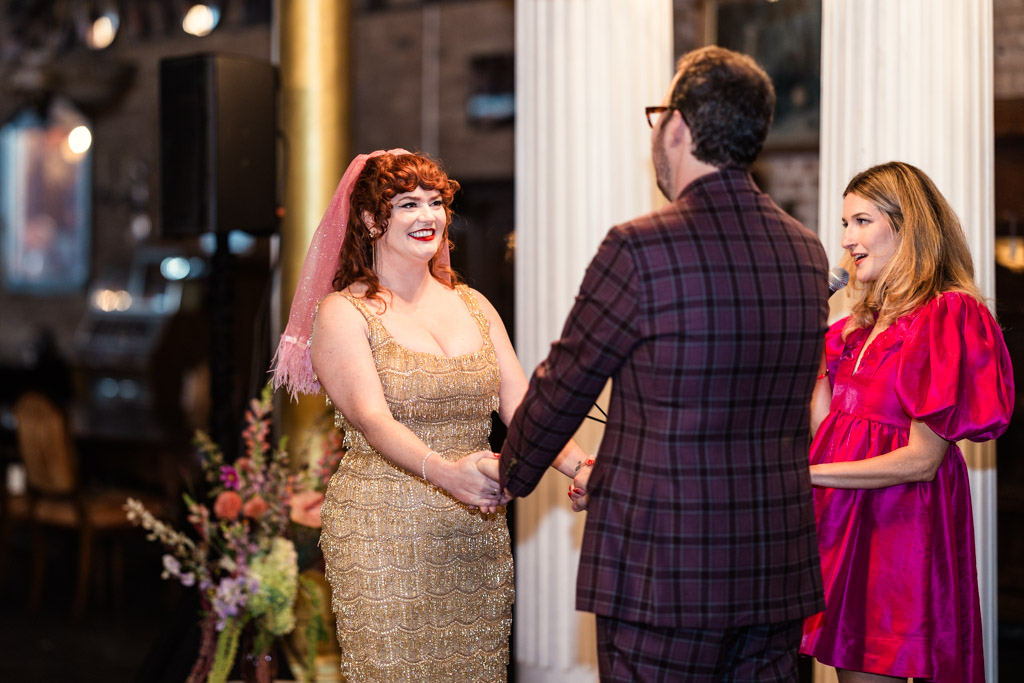 Bride smiles while holding hands with the groom at their Salvage One wedding ceremony, with an officiant in a bright pink dress standing nearby