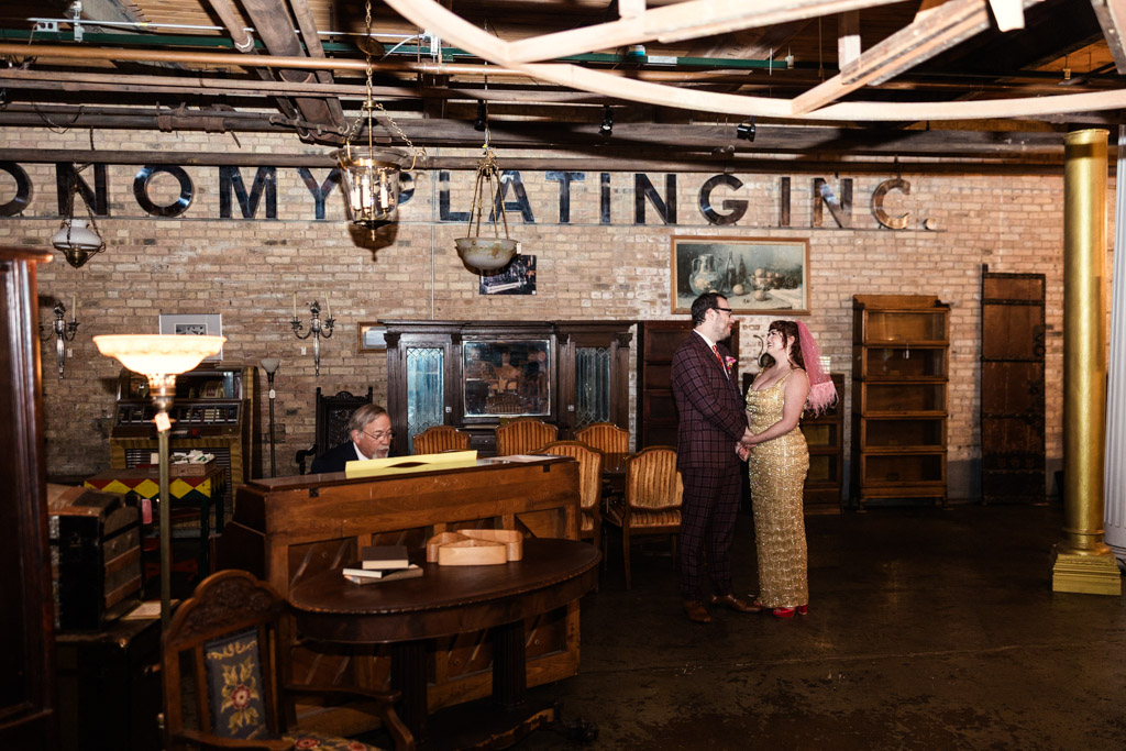 Bride and groom dressed in vintage clothes stand together holding hands during their Salvage One wedding ceremony, while a man plays piano nearby