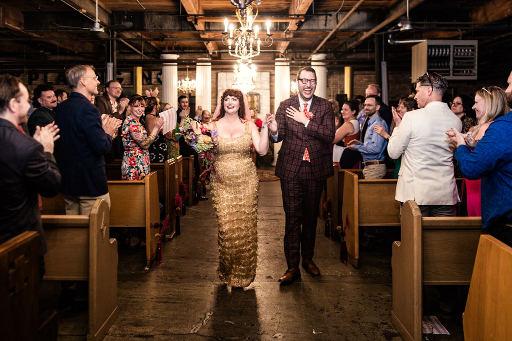 Newlyweds walks down the aisle at their Salvage One wedding as guests stand and clap in a warmly lit venue.