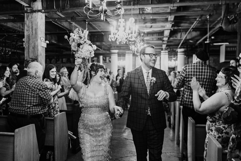 Black and white photo of happy bride and groom walking down the aisle holding hands as guests cheer and applaud after their Salvage One wedding ceremony