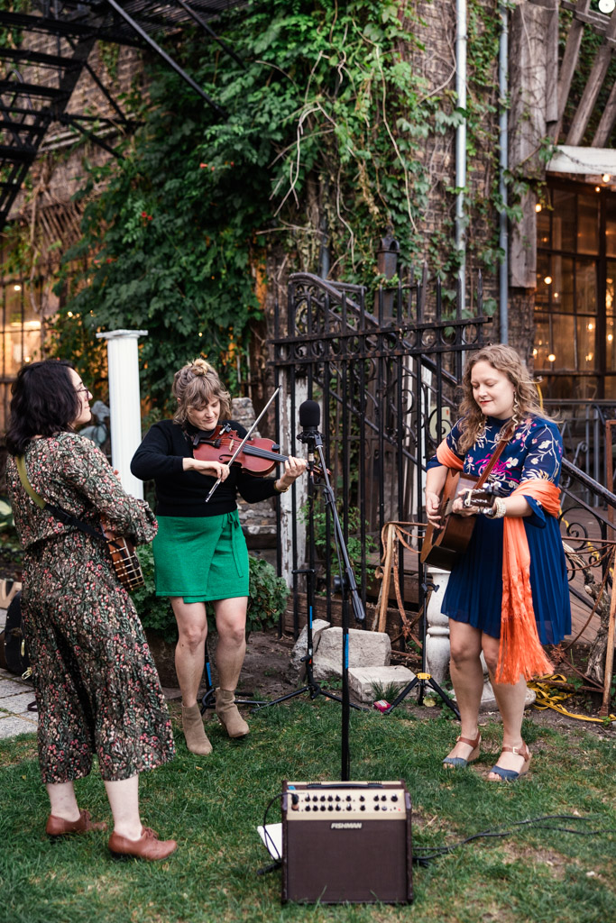 Live musicians play banjo, guitar and violin outdoors surrounded by greenery and rustic decor during Salvage One wedding reception