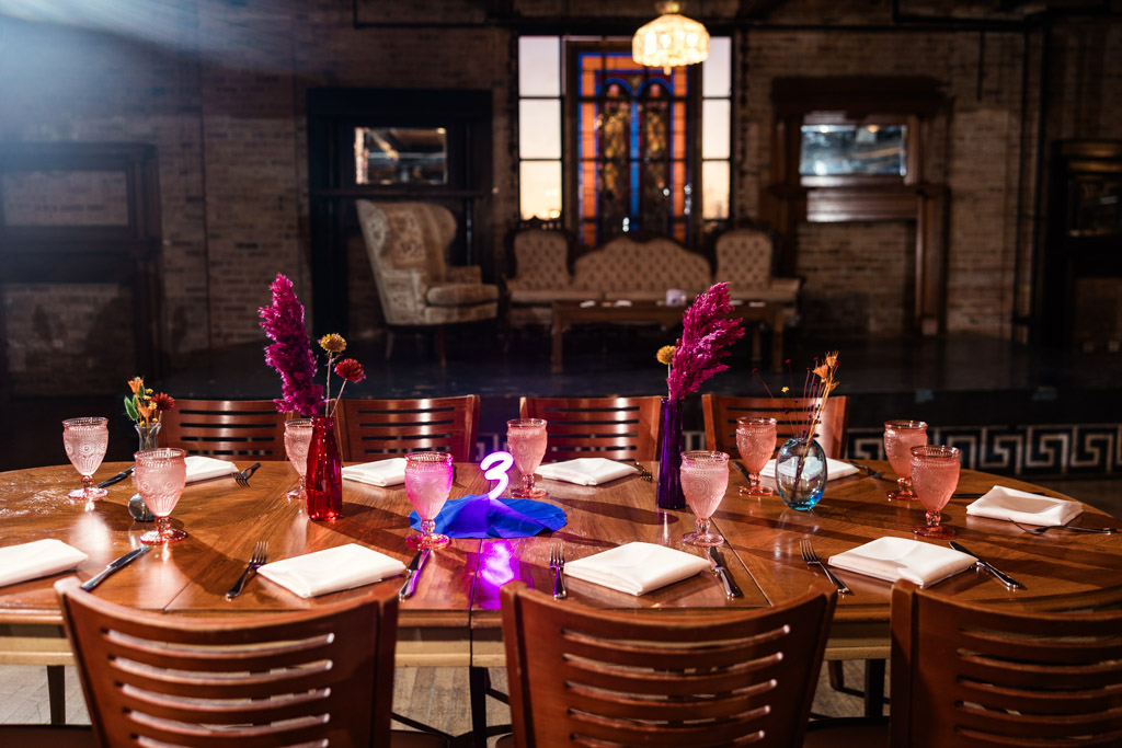 Guest table with pink drinks, flowers, and napkins, set in a vintage room with a stage and sofas or Salvage One wedding celebration